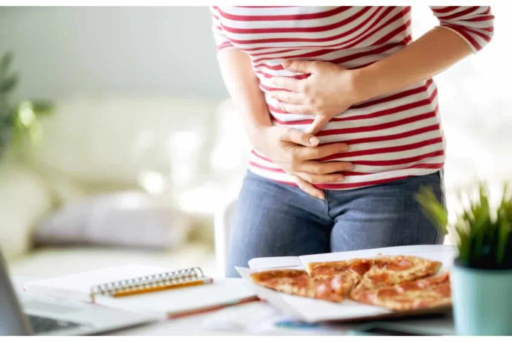 une personne se tient le ventre avec ses deux mains. elle est debout, devant une table sur laquelle on devine des parts de pizza, un calepin et un crayon ainsi qu'une plante.